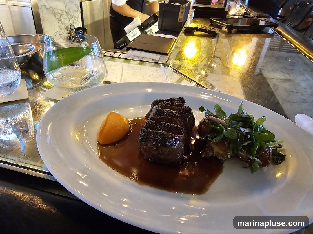 A plate of sliced steak with a side of potatoes and greens, served with a slice of orange and a rich sauce. Two glasses of water and a cocktail with a leaf garnish are visible on the bar. The background shows a bar area with a bartender in uniform.