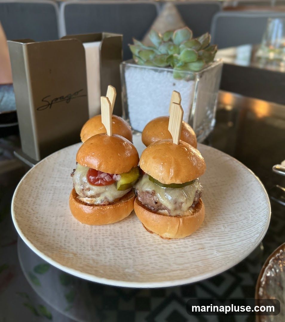 Four sliders are presented on a textured white plate. The sliders have brioche buns, beef patties, melted cheese, pickles, and what appears to be chopped onions and ketchup. In the background, a decorative planter with a succulent and a branded napkin holder with the name "Spago" are visible. The setting appears to be a restaurant or dining establishment.