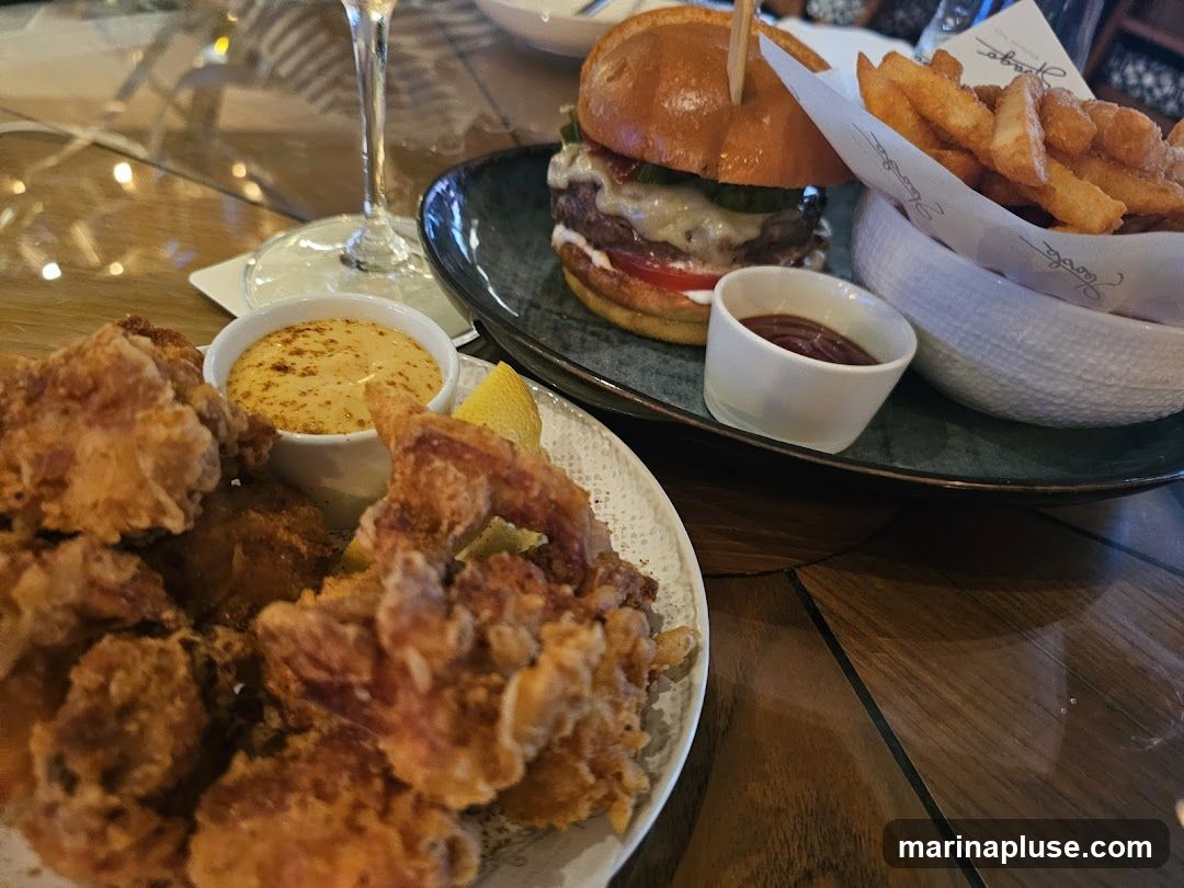 A close-up shot of a table setting with two plates of food. In the foreground, a plate of golden-brown fried chicken pieces is served with a small bowl of dipping sauce and a lemon wedge. In the background, a gourmet burger with melted cheese, lettuce, and tomato is presented on a dark plate alongside a portion of thick-cut fries and a small dish of ketchup. A wine glass is also visible on the table.