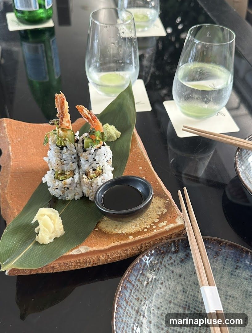 A close-up shot of a sushi roll served on a leaf with wasabi, pickled ginger, and soy sauce. There are also glasses of water with a green liquid at the bottom and chopsticks on the table.