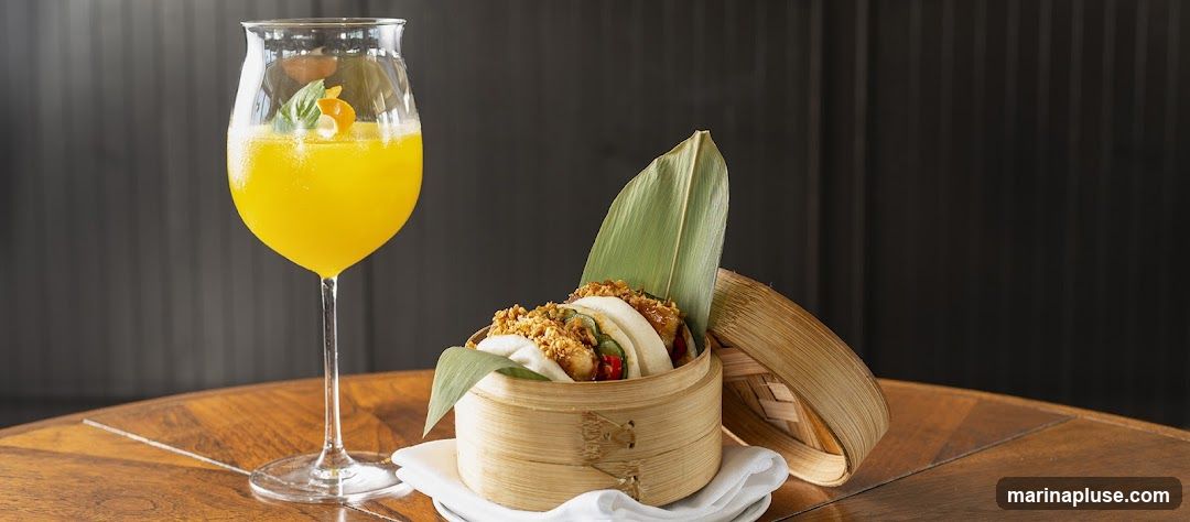 A close-up shot of a bamboo steamer filled with bao buns and a tall glass of a bright orange cocktail. The bao buns appear to be filled with a savory mixture and topped with crispy bits and greens. The cocktail is garnished with a sprig of basil and a twist of orange peel. The food and drink are presented on a wooden table against a dark, textured background.