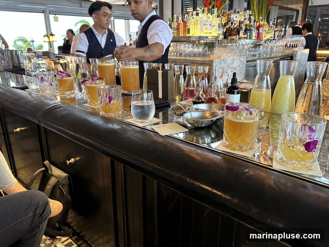 A bar counter with several cocktails and glasses of water. Bartenders in uniform are visible in the background, preparing drinks. The bar is well-stocked with bottles of liquor and mixers.