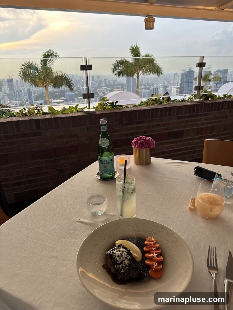 A table set for a meal on a rooftop terrace overlooking a city skyline at dusk. The table is covered with a white tablecloth and set with plates of food, drinks, and cutlery. A bottle of San Pellegrino water is on the table, along with a cocktail and a glass of water. A plate of food, appearing to be a piece of meat with a lemon wedge and sliced carrots, is in the foreground. In the background, palm trees and city buildings are visible under a cloudy sky.
