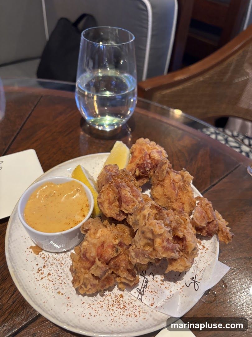 A plate of crispy fried chicken pieces with a side of dipping sauce and lemon wedges sits on a wooden table. A glass of water is also visible.