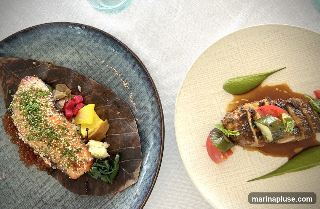 Two plates of food are presented on a white tablecloth. The plate on the left contains a piece of salmon, garnished with sesame seeds and chives, served on a large leaf with rice, pickled vegetables, and roe. The plate on the right features sliced grilled meat with a dark sauce, accompanied by tomatoes, cucumbers, and a green puree.