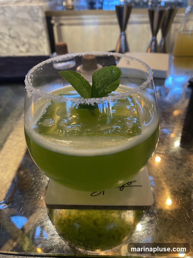 A close-up shot of a vibrant green cocktail in a stemmed glass. The rim of the glass is coated with salt. The drink is garnished with fresh mint leaves and contains a large ice cube. The cocktail is placed on a coaster with a signature in cursive. The background appears to be a bar area with bottles and bar tools visible.