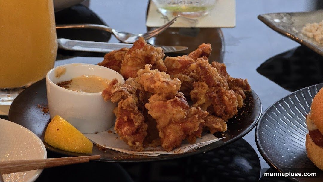 A close-up shot of a plate of crispy fried chicken bites, served with a small bowl of dipping sauce and a lemon wedge. A glass of orange juice and part of a burger are also visible on the table.