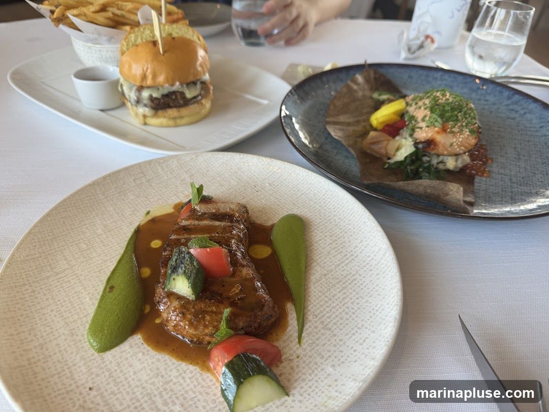 A close-up view of three elegantly plated dishes on a white tablecloth. In the foreground, a slice of meat is served with a rich brown sauce, vibrant green puree, and garnished with tomato and cucumber. To the left, a gourmet burger is presented with a side of french fries and a small dipping sauce. In the background, a piece of salmon is artfully arranged with vegetables on a dark blue plate. Glasses of water are visible on the table.