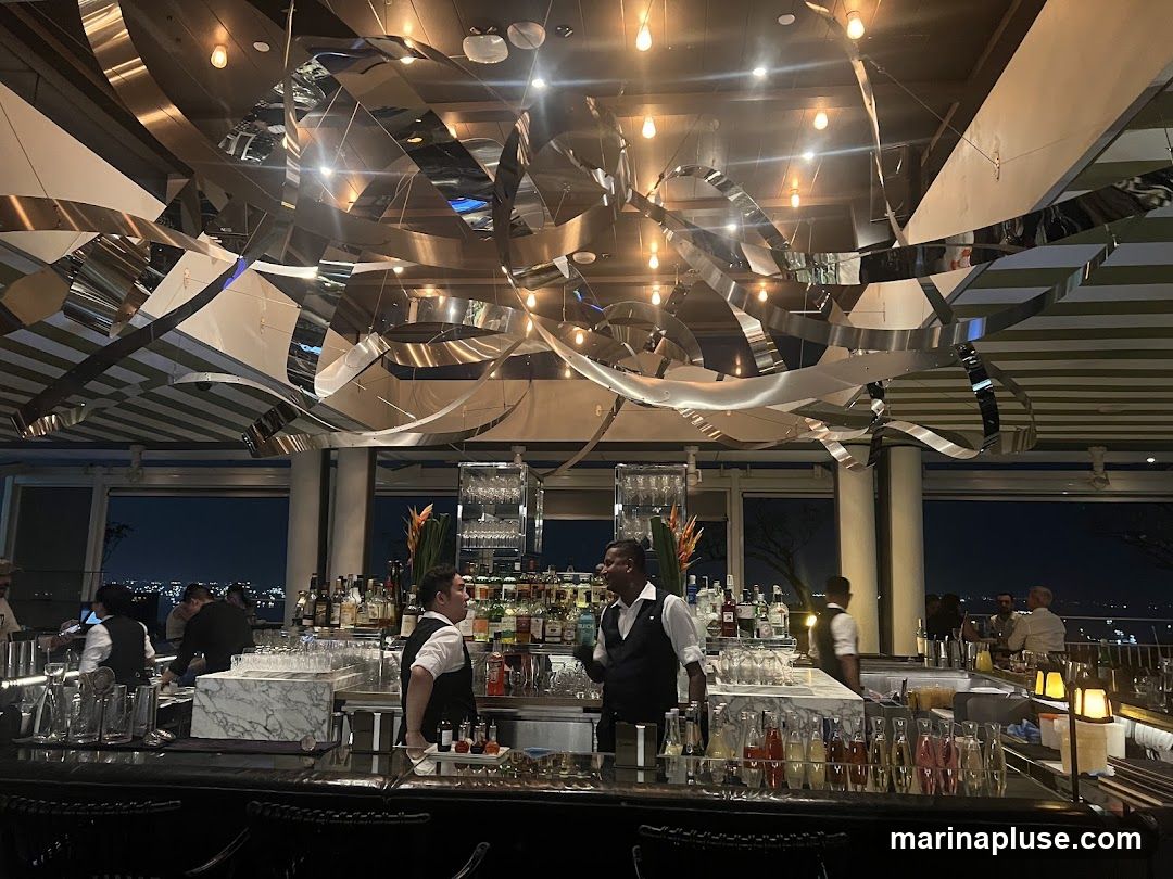 The interior of a bar with a view of the city at night. Bartenders are visible behind a well-stocked bar, preparing drinks. The ceiling features a modern, abstract metallic sculpture and striped panels.