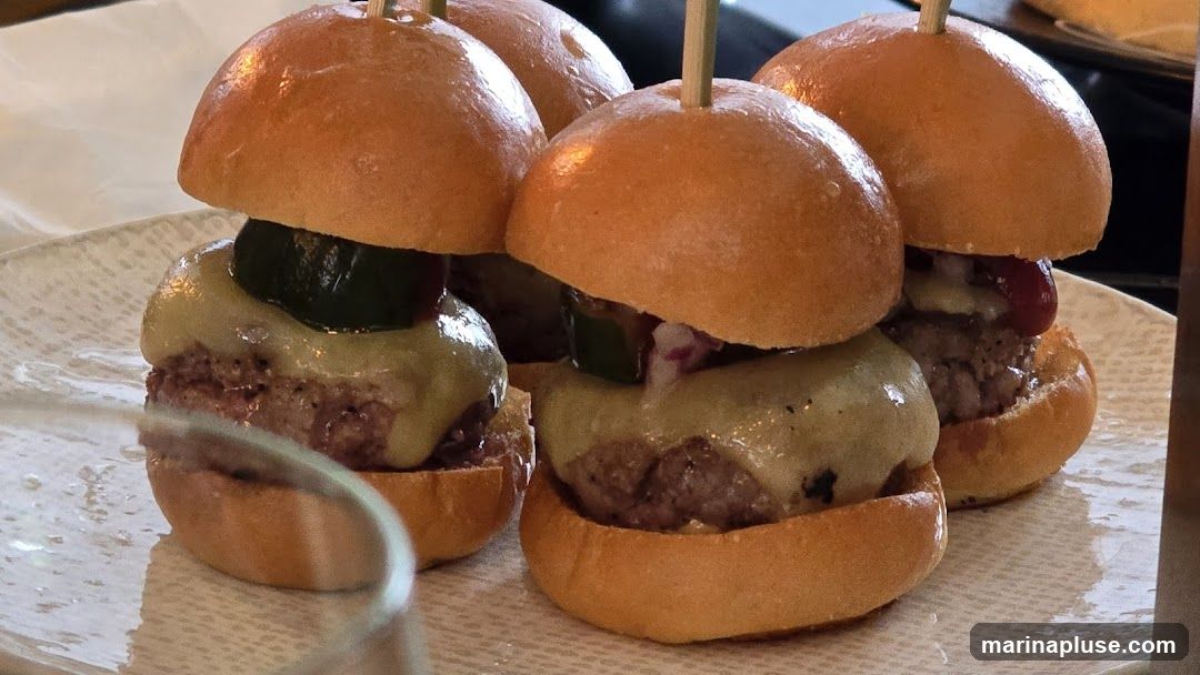A close-up shot of four sliders on a textured plate. Each slider has a toasted bun, a beef patty, melted cheese, and a pickle slice. Some sliders also appear to have red onion and ketchup.