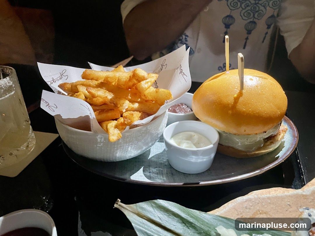 A close-up shot of a burger and fries served at a restaurant. The burger is on a plate with two small dipping sauces, and the fries are in a white bowl lined with paper that has a logo. A drink with a salted rim is visible to the left.