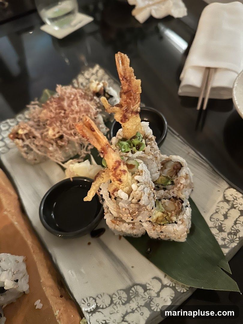 A close-up view of a sushi platter featuring several pieces of sushi rolls topped with tempura shrimp and a side of soy sauce. Another dish, possibly tempura, is visible in the background. The setting appears to be a restaurant with chopsticks and a napkin on the table.