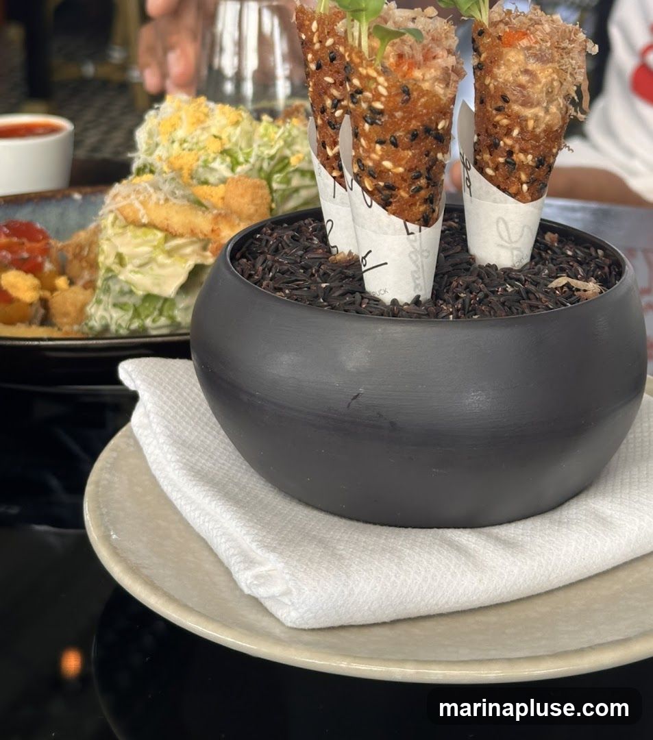 Three crispy cones filled with a savory mixture and topped with microgreens are presented in a black bowl filled with black rice. In the background, a Caesar salad with croutons and a small dish of sauce are visible.