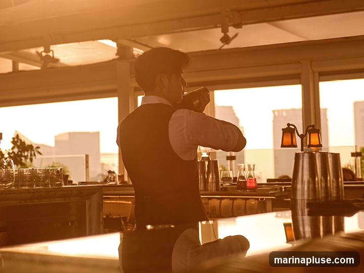 A bartender, silhouetted against a warm, golden sunset, is seen from behind as they prepare a drink at a bar. They are wearing a vest over a white shirt and are actively shaking a cocktail shaker. The bar is equipped with various bar tools, shakers, glasses, and bottles of colorful liquids. In the background, large windows offer a view of city buildings, suggesting the bar is located in an urban setting, possibly on a rooftop. Decorative lamps with amber light add to the ambiance.