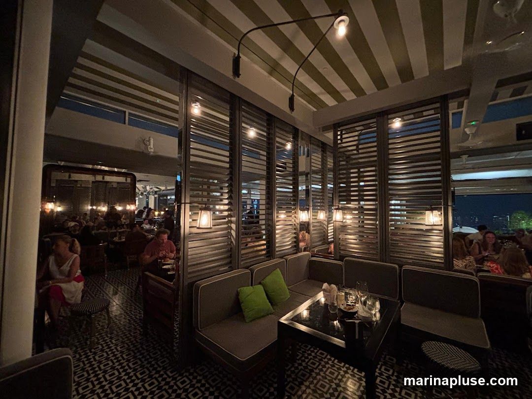 A dimly lit restaurant interior with patterned tile flooring and striped ceiling. Booth seating with green cushions is arranged around a dark table set with glasses and small dishes. In the background, other diners are seated at tables, and large slatted windows offer a view of the city at night.