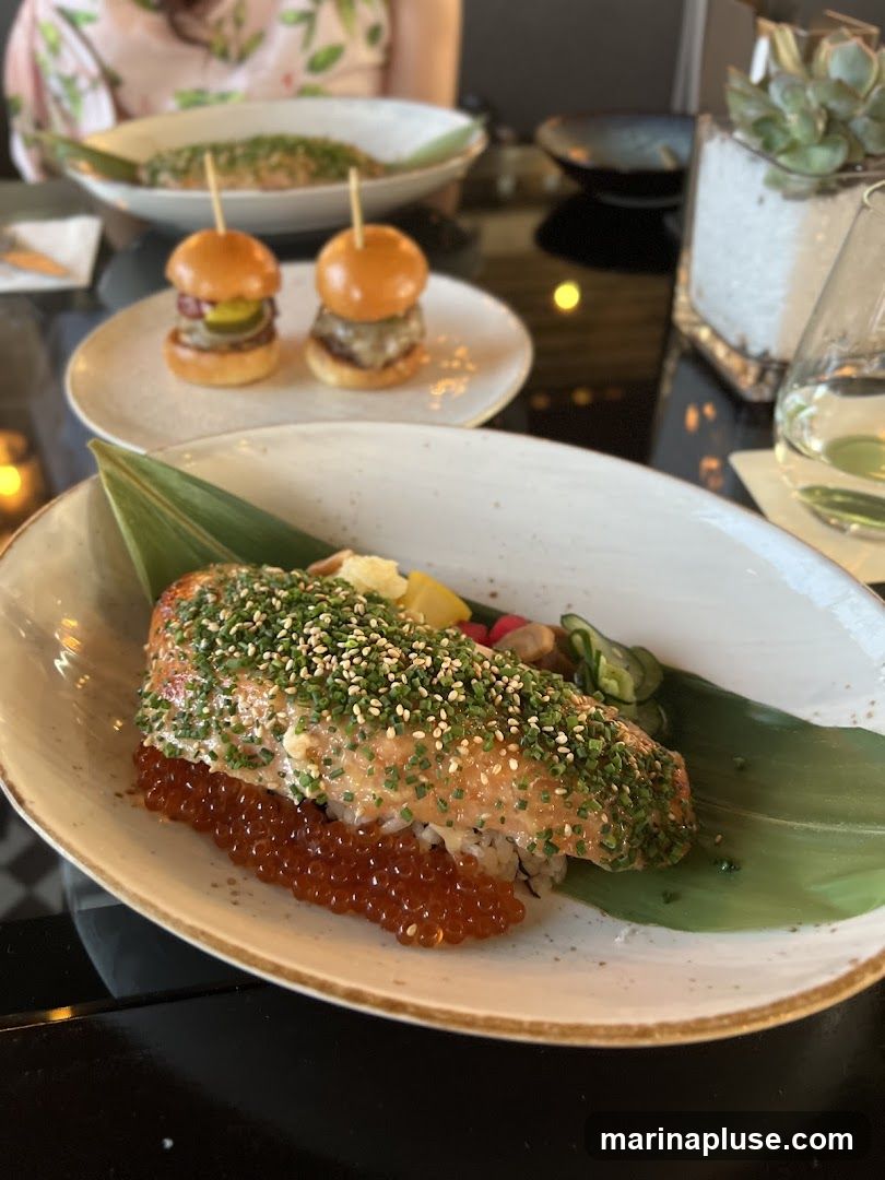 A close-up of a salmon dish served over rice with ikura (salmon roe) and garnished with sesame seeds and chives. The dish is presented on a large green leaf. In the background, two mini burgers are visible on a plate, and another dish with what appears to be a fish fillet is out of focus. A glass of water and a potted succulent are also on the table.