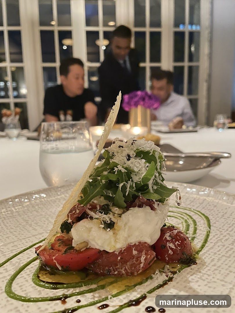 A close-up of a gourmet salad featuring fresh mozzarella, roasted tomatoes, arugula, and a crispy cracker, artfully arranged on a textured white plate. The dish is garnished with grated cheese and a vibrant green sauce, with balsamic glaze drizzled around the edges. In the background, blurred figures of diners and a server are visible in a softly lit dining room with large windows.