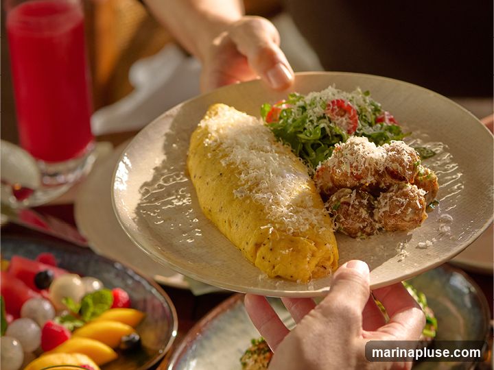 A close-up shot of a person's hands holding a plate with an omelette, a side of roasted potatoes, and a small salad. The omelette is sprinkled with grated cheese. A glass of red juice and a bowl of fruit salad are visible in the background.