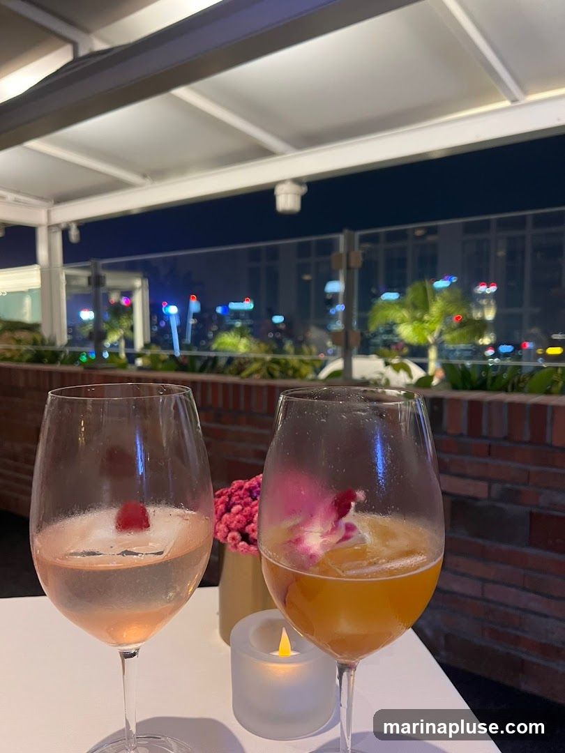 Two wine glasses filled with colorful cocktails sit on a white table. One glass contains a pale pink drink with a raspberry and ice, while the other holds an amber-colored beverage with ice and a flower garnish. A small, lit candle and a vase of pink flowers are also on the table. In the background, a blurred cityscape at night is visible through a glass railing, with palm trees and city lights suggesting a tropical or urban rooftop setting.