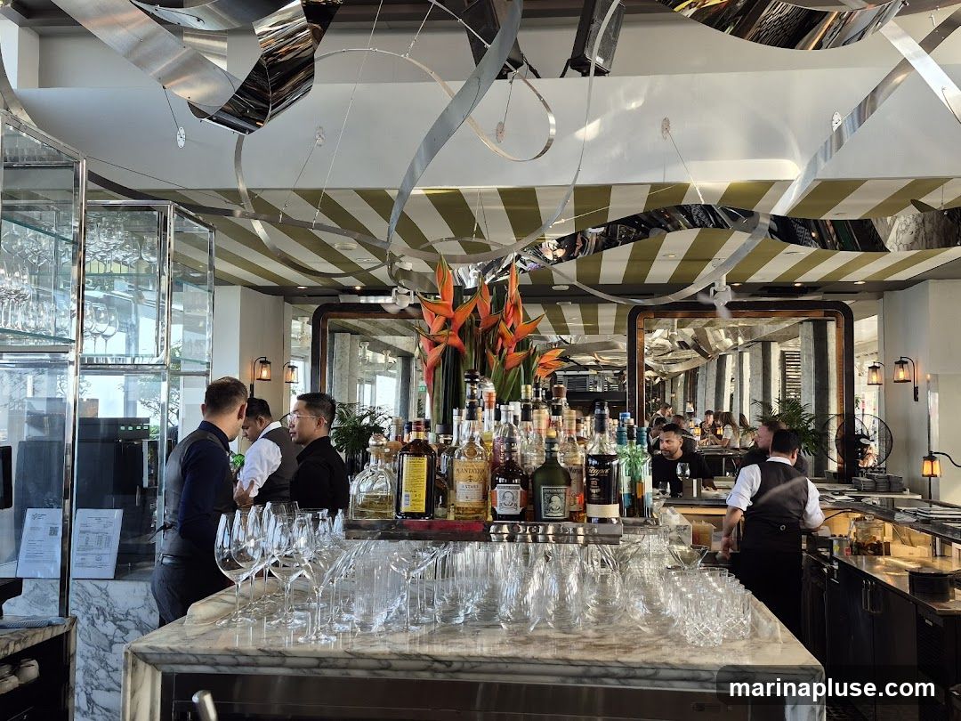 A well-lit bar interior with a marble countertop in the foreground, lined with numerous wine glasses. Behind the counter, a variety of liquor bottles are displayed on shelves. Several staff members are visible, some interacting with customers and others working behind the bar. The ceiling features a striking design with metallic ribbons and striped awnings. Mirrors reflect the scene, adding depth to the space.