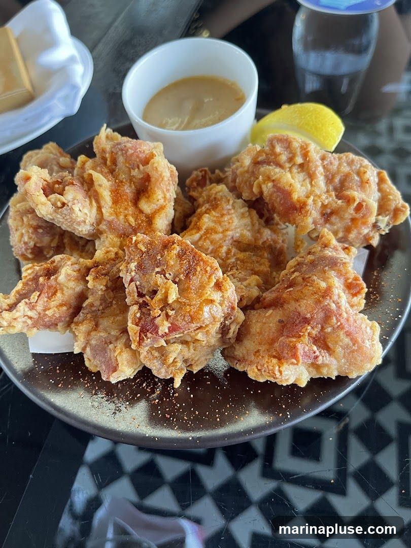 A plate of golden-brown fried chicken bites is served with a small bowl of dipping sauce and a lemon wedge. The chicken appears crispy and seasoned. The plate is on a table with a black and white geometric pattern.