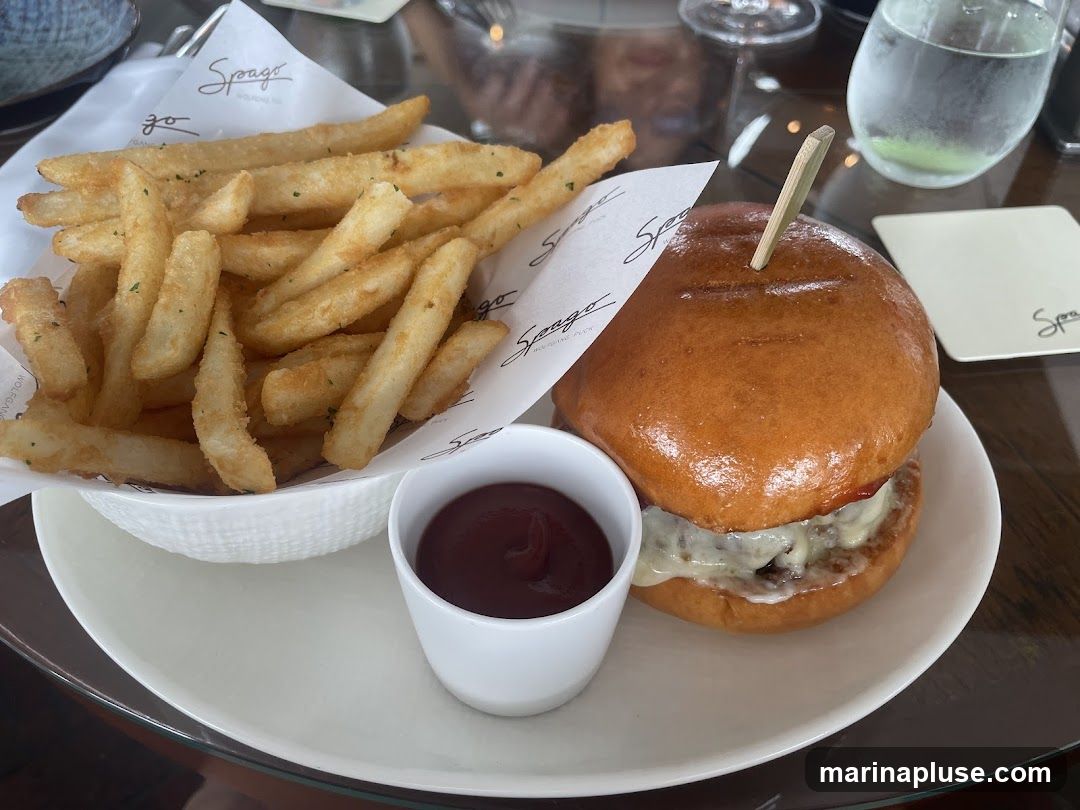 A close-up shot of a burger and fries served on a white plate. The burger has a glossy bun and appears to have cheese and a patty inside. A small white ramekin of ketchup is placed next to the burger. A white bowl filled with golden french fries is on the left side of the plate. The fries are seasoned with herbs. The food is presented on a table with a glass of water and a coaster visible in the background. The branding "Spago" is visible on the paper lining the fries and on the coaster.