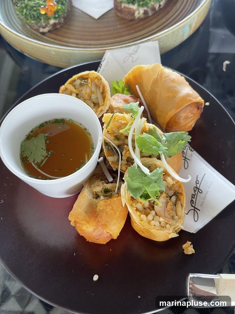 A plate of golden-brown spring rolls, some whole and some cut in half to reveal a filling of rice and meat, are arranged around a small white bowl of dipping sauce. The spring rolls are garnished with fresh cilantro and thinly sliced green onions. A white napkin with a logo and text is tucked beneath the spring rolls. In the background, out of focus, are two small appetizers topped with green herbs and red roe.