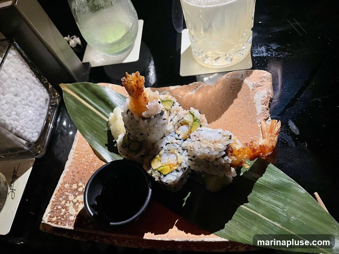 A plate of sushi rolls with tempura shrimp and avocado is served on a leaf with a small dish of soy sauce. Two cocktails are visible in the background, one with ice and mint, the other in a faceted glass. A container of white pebbles is also in the frame.