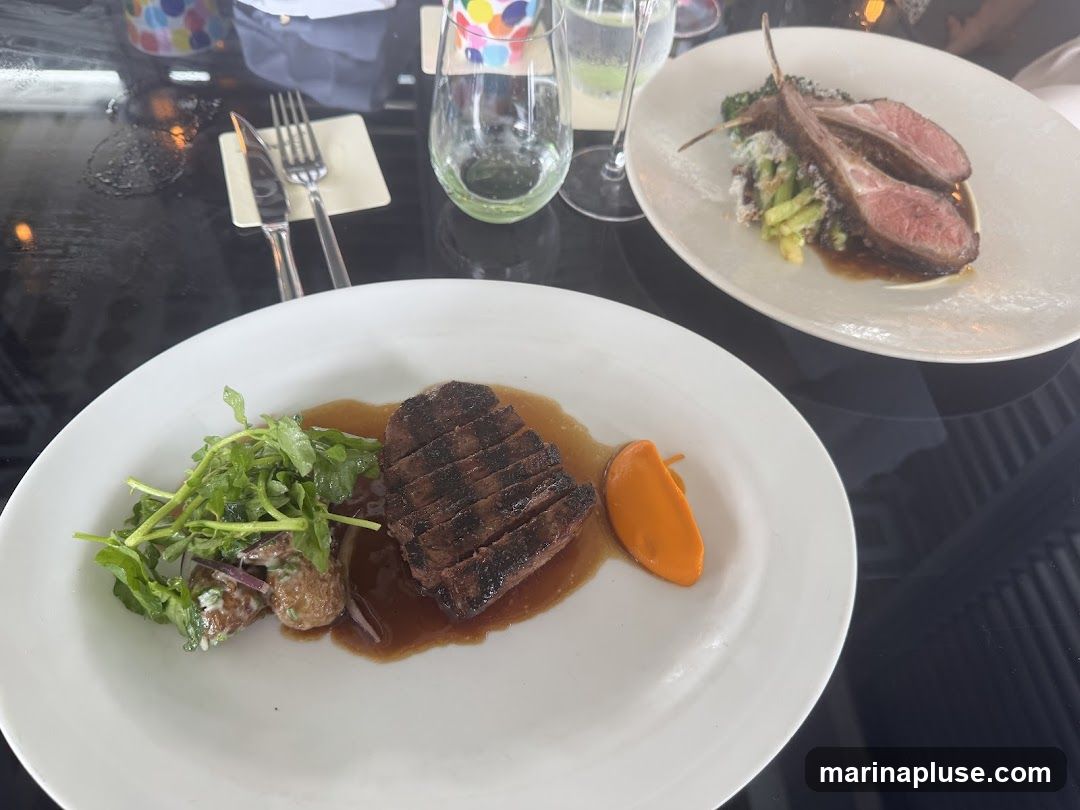 A close-up shot of two white plates, each containing a meat dish. The plate in the foreground features sliced steak with a side of potato salad and a dollop of orange puree. The plate in the background displays lamb chops served with a green vegetable medley. Cutlery and glasses are visible on the dark, reflective table.