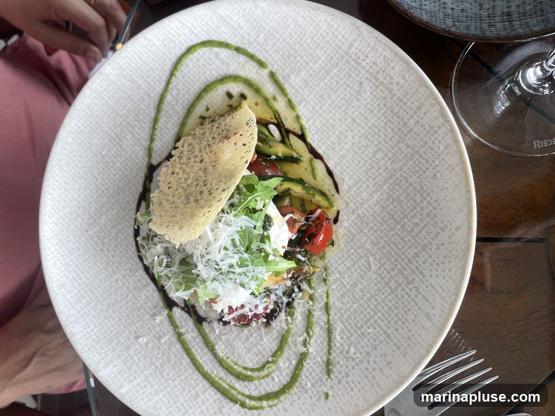 A close-up overhead shot of a gourmet salad served on a textured white plate. The salad features thinly sliced zucchini, cherry tomatoes, arugula, and a dollop of white cheese, possibly mozzarella or burrata. It is garnished with grated Parmesan cheese, a crispy cheese crisp, and drizzled with a vibrant green sauce and a dark balsamic glaze. A fork is visible in the lower right corner, and a wine glass is partially visible in the upper right. The setting appears to be a restaurant.