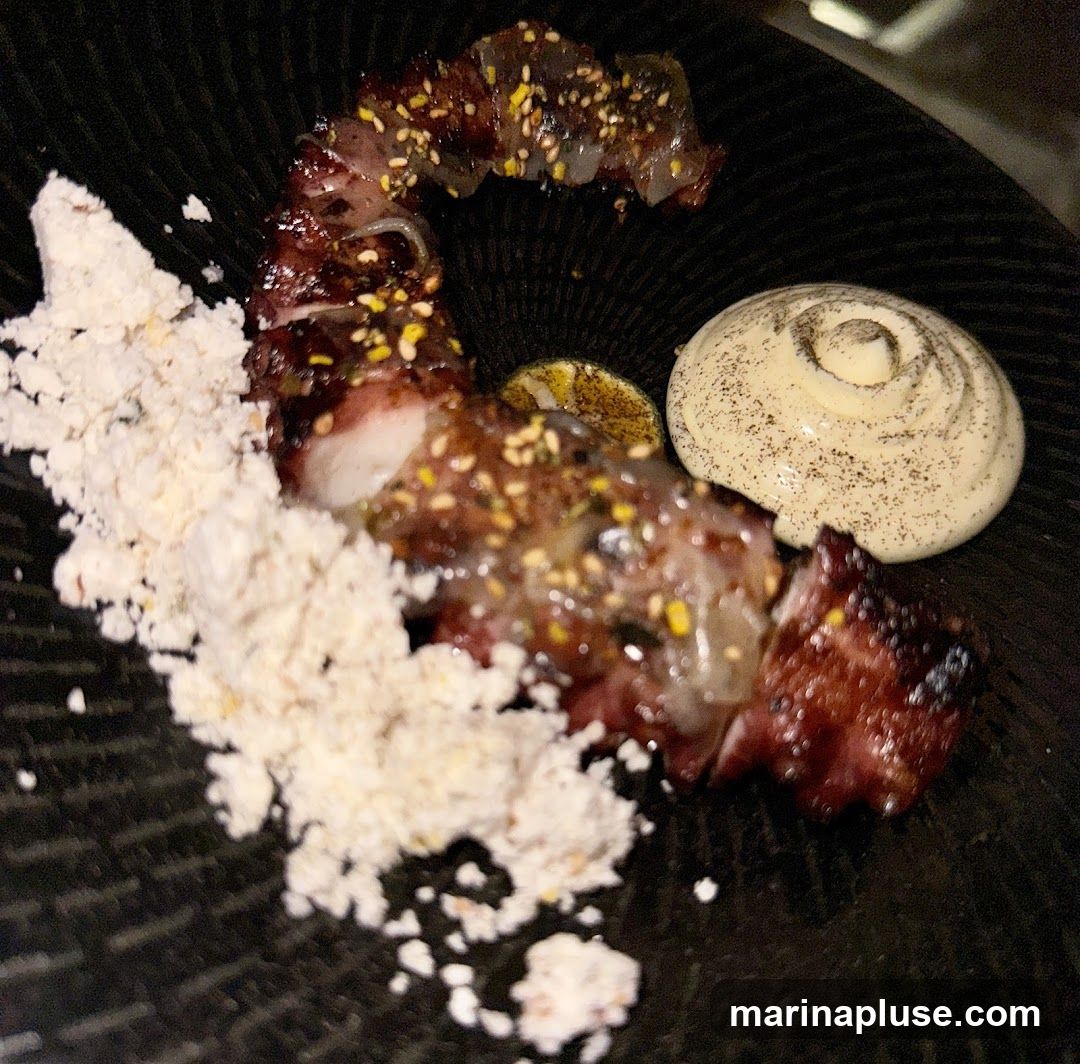 A close-up shot of a gourmet dish served on a black plate. The dish features pieces of grilled meat or seafood, glazed with a dark sauce and sprinkled with sesame seeds and yellow zest. A dollop of creamy sauce, possibly aioli or a puree, is swirled on the side, dusted with black pepper. A slice of grilled lemon or lime is also visible. A pile of white, powdery substance, perhaps a crumble or a foam, is placed on the left side of the plate. The lighting is dim, highlighting the textures and colors of the food.