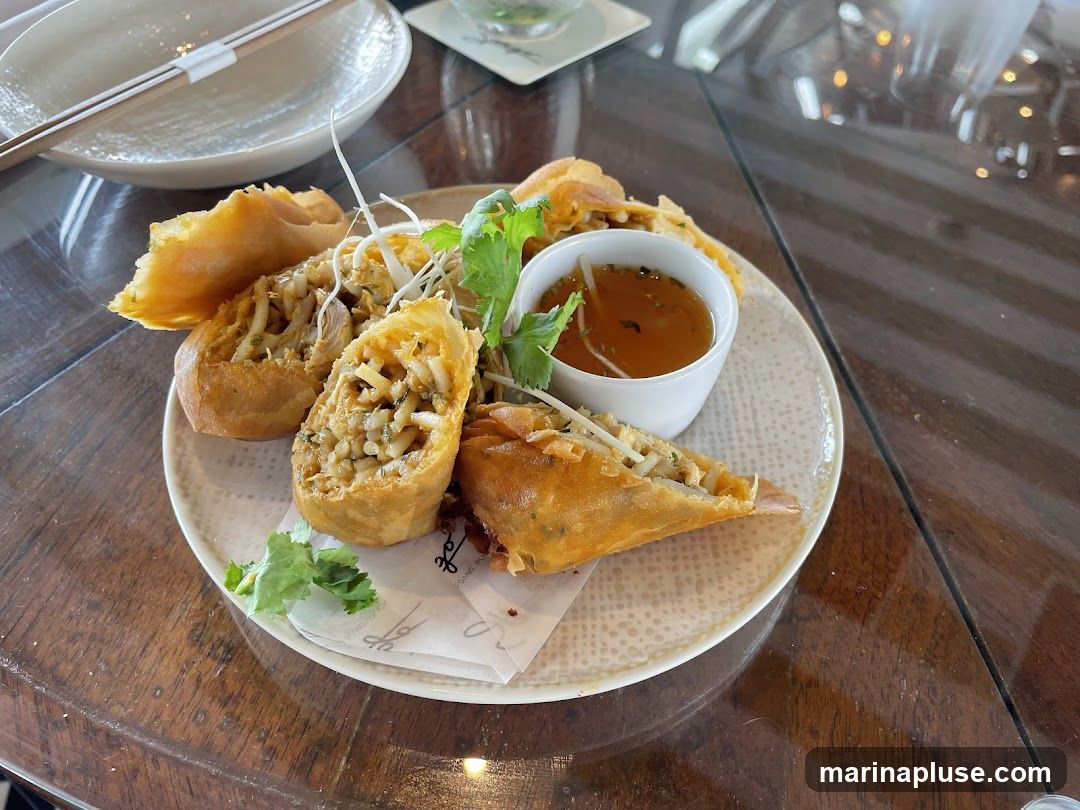 A plate of golden-brown fried spring rolls, some whole and some cut in half to reveal a filling of shredded vegetables and possibly meat. A small white bowl of dipping sauce, garnished with cilantro, is placed on the plate. The plate is set on a wooden table, with chopsticks and a white bowl visible in the background. A small piece of paper with a logo is under the spring rolls.