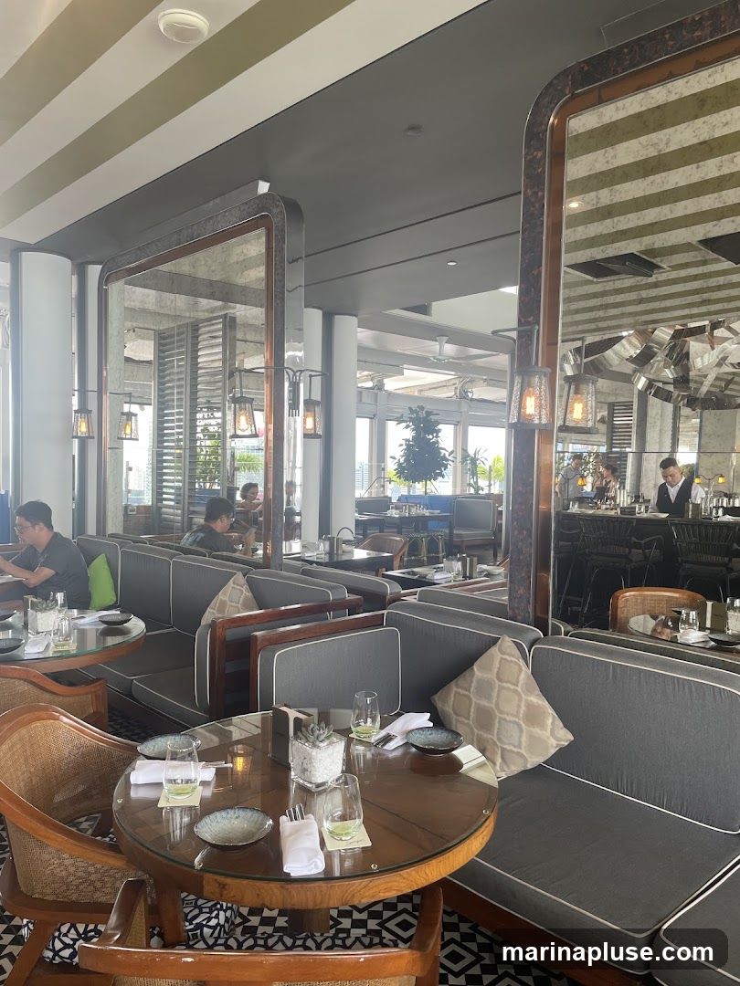 A view of a restaurant interior with tables and seating. The tables are set with plates, glasses, and napkins. There are large mirrors on the walls, and pendant lights hang from the ceiling. Several patrons are seated at tables, and staff can be seen behind the bar.