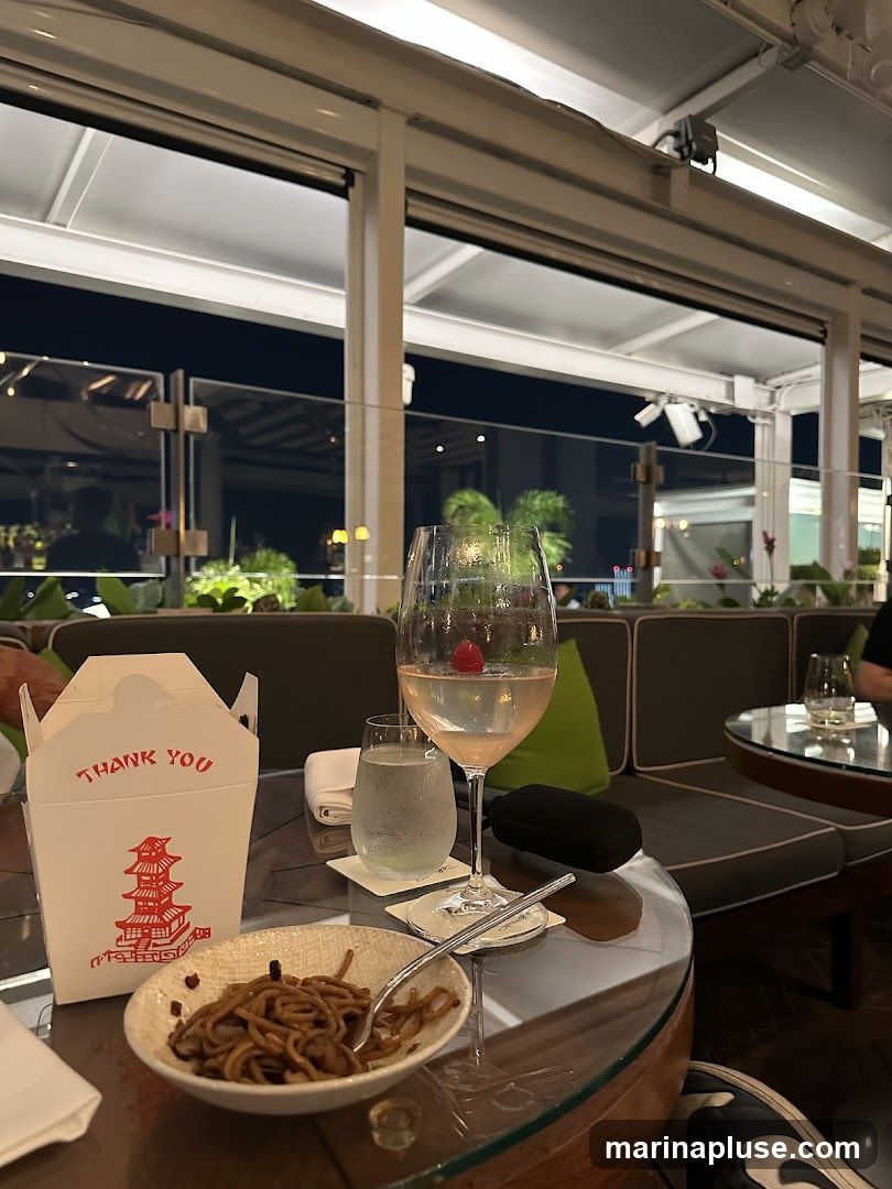 A table setting on a rooftop patio at night, featuring a glass of rosé with a raspberry, a glass of water, a small bowl of noodles, and a takeout container with "THANK YOU" printed on it. The background shows a blurred view of city lights and greenery, with comfortable seating and overhead lighting.