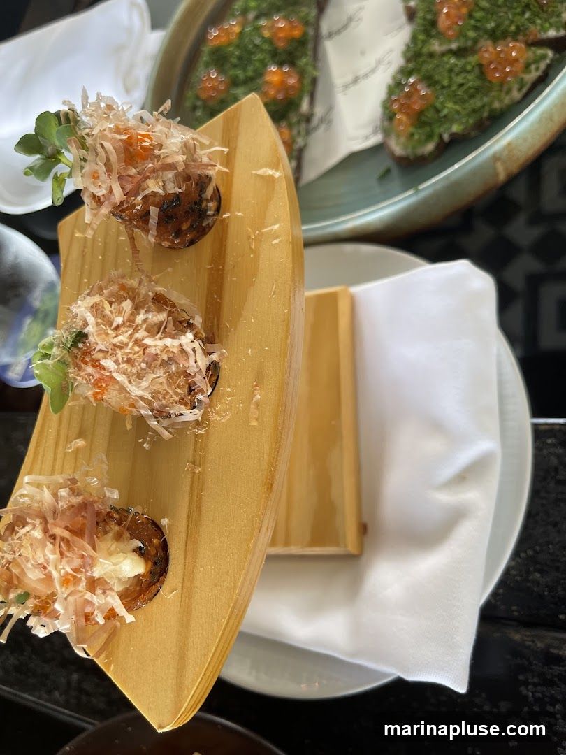 Three small, round appetizers are presented on a curved wooden board. Each appetizer is topped with shredded bonito flakes, a dollop of orange roe, and a sprig of green garnish. In the background, another dish holds several pieces of toast covered in green herbs and orange roe.
