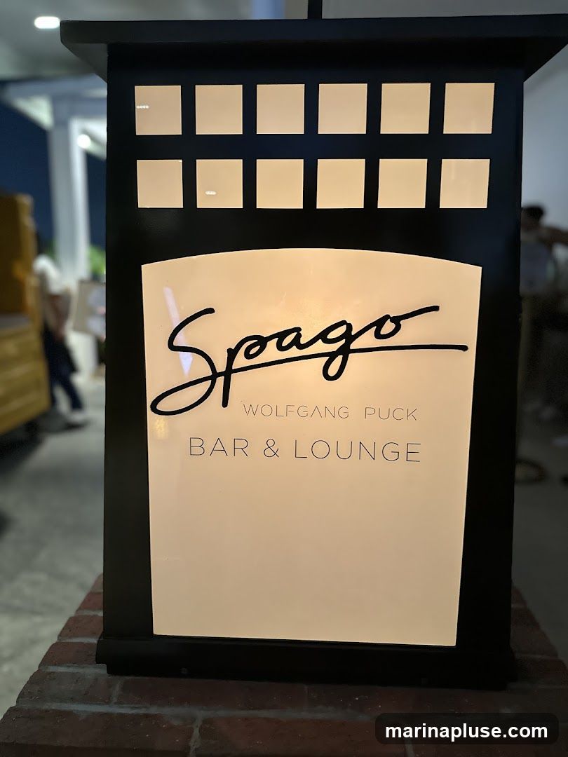 A close-up shot of a lit sign for Spago Wolfgang Puck Bar & Lounge. The sign is black with a white illuminated panel. The Spago logo is in a stylized black script at the top of the panel, with "WOLFGANG PUCK" and "BAR & LOUNGE" printed below in a simple font. The sign is mounted on a brick base. In the blurred background, people can be seen walking.