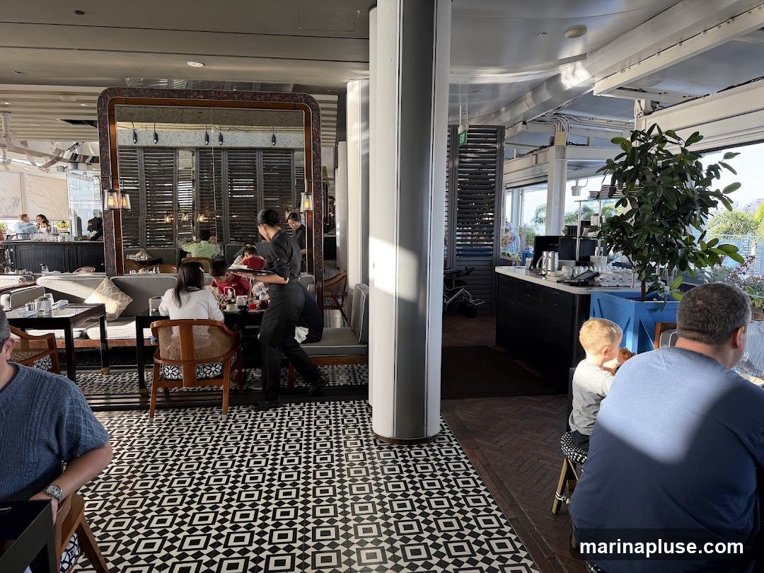 A view of a restaurant interior with patrons seated at tables. A server is seen carrying a tray. The floor has a distinctive black and white geometric tile pattern. Sunlight streams in from the right, illuminating a potted plant and a counter area.