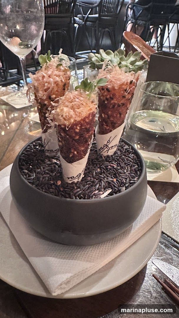 Three appetizer cones filled with a savory mixture and topped with shredded ingredients and microgreens, presented in a bowl of black rice. The cones are wrapped in paper with a logo visible.