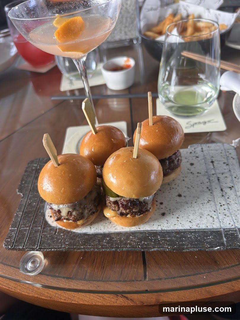 A close-up shot of four sliders on a textured platter, served with toothpicks. In the background, a cocktail with an orange peel garnish is visible, along with a glass of white wine and a bowl of french fries. The setting appears to be a restaurant or bar.