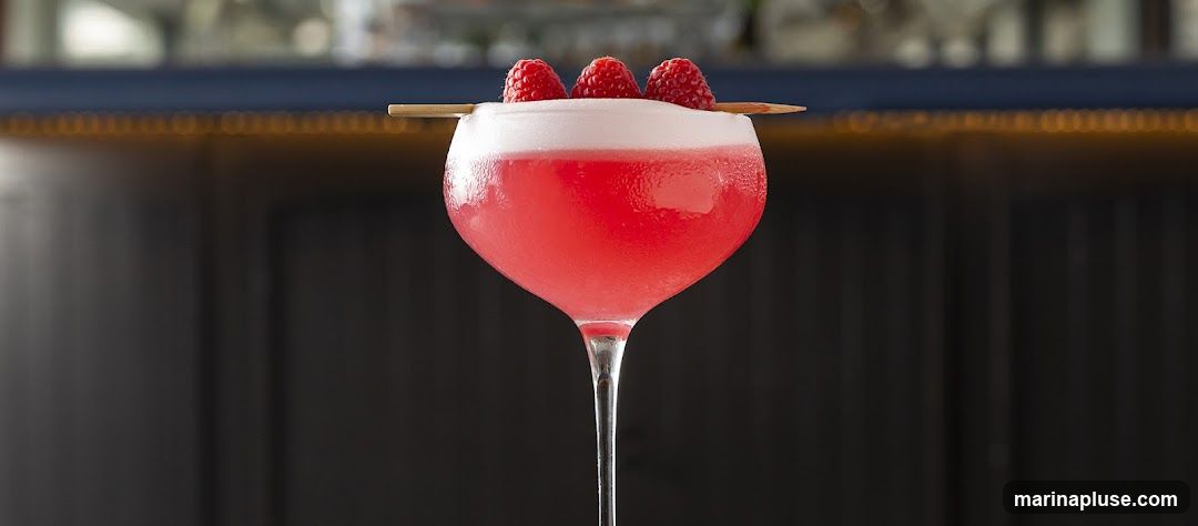 A close-up shot of a pink cocktail in a coupe glass, garnished with three fresh raspberries on a skewer. The drink has a frothy top and condensation on the glass. The background is blurred, showing a dark, vertical-lined surface and a dark blue bar with warm lighting.