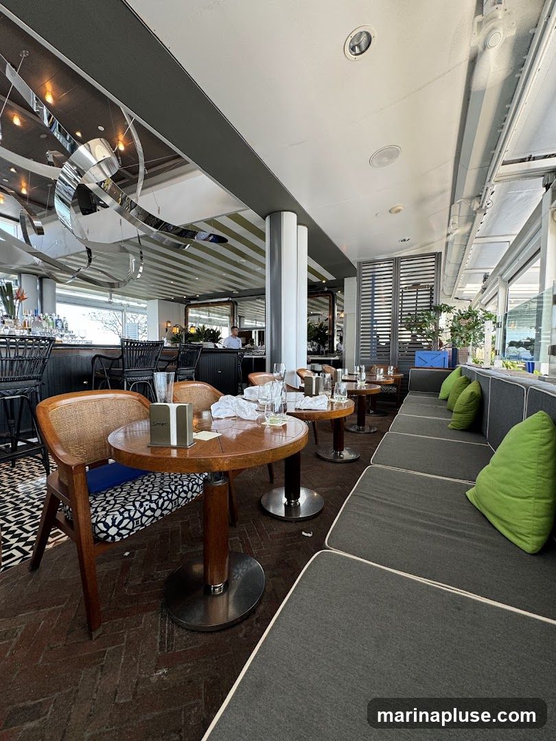 The interior of a restaurant with tables and seating. There is a bar area with bottles of alcohol visible. The seating includes chairs and a long banquette with green cushions. The decor is modern with a large abstract sculpture hanging from the ceiling.
