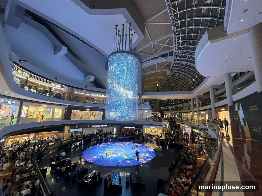 The image shows the interior of a multi-level shopping mall. A large, cylindrical light installation hangs from the ceiling, and a circular water feature with a projected light show is visible in the center of the ground floor. Shops such as FILA KIDS, SO PHO, PUTIEN, RAMEN IPPUDO, and STARBUCKS are visible on the upper levels. Many people are browsing and seated around the water feature. The architecture features a glass ceiling and curved balconies.