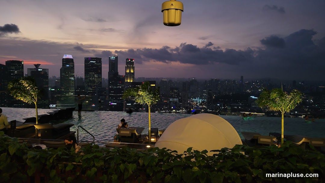 An infinity pool overlooks a city skyline at dusk. The sky is a deep purple with hints of orange and pink. Silhouetted skyscrapers are visible in the distance, illuminated by city lights. Several trees are planted around the pool, and lounge chairs are set up for relaxation. A white cabana-like structure is also visible. People are seen in the pool and seated at tables. A golden light fixture hangs from above.