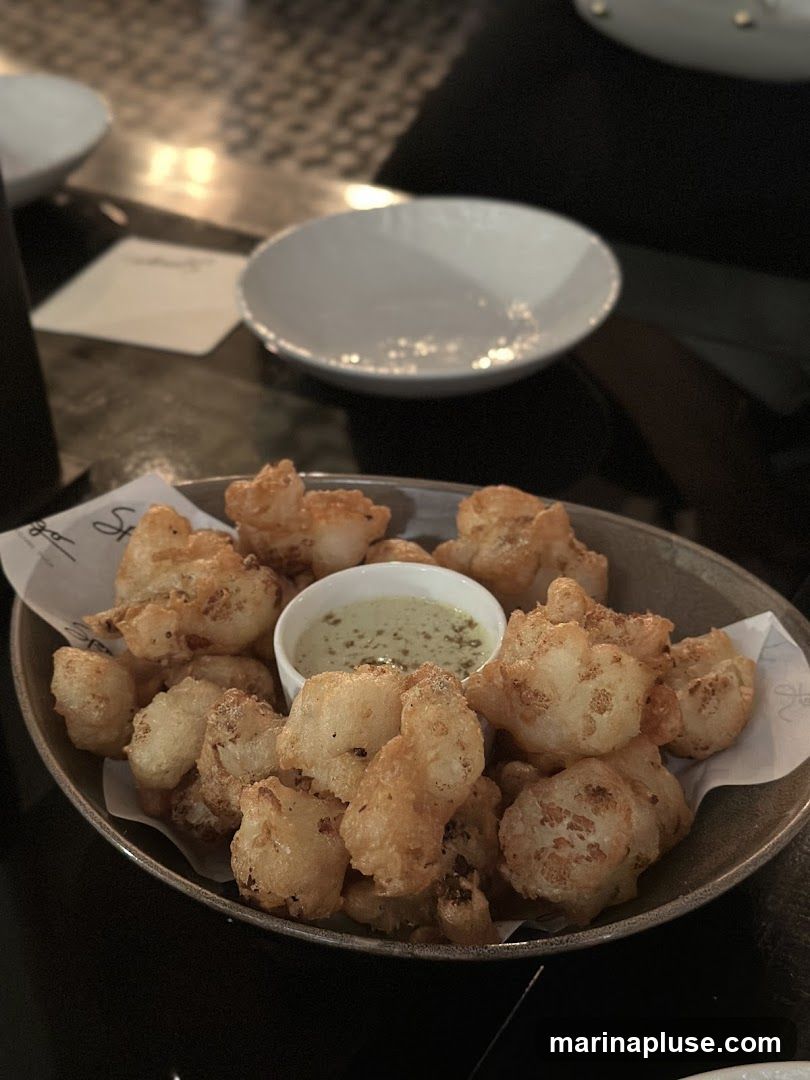 A close-up shot of a bowl filled with golden-brown fried cauliflower bites, served with a small dish of creamy green dipping sauce. The cauliflower is lightly battered and appears crispy. The dish is presented on a dark surface, with blurred white bowls and a napkin in the background, suggesting a restaurant setting.