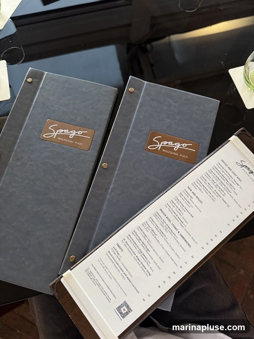 A menu from Spago by Wolfgang Puck, featuring sections for Snacks and Light Bites, Raw and Rolled, Bigger Bites, Steaks & Sandwiches, and Sweets.