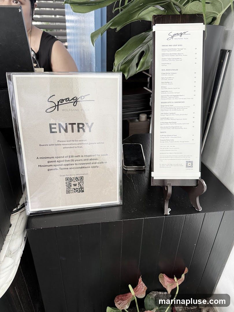 A sign at the entrance of Spago restaurant indicates that guests should wait to be seated and that a minimum spend of $30 nett is required per guest aged five and above. A menu is displayed next to the sign, listing various food and drink options.