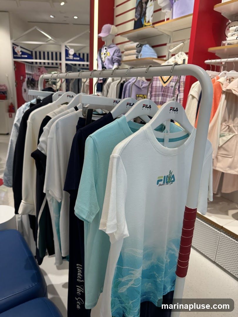 A rack of children's t-shirts and pants hangs in a store. The t-shirts are various colors including white, navy, and light blue, with some featuring graphics. The pants are dark blue with white lettering that reads "Under The Sea". In the background, mannequins are dressed in children's clothing, and shelves display more apparel. The store has red and white accents.