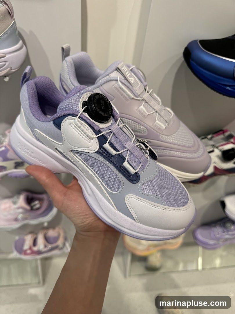 A hand holds up a pair of purple and white Fila sneakers with a BOA lacing system. Other shoes are visible on shelves in the background.