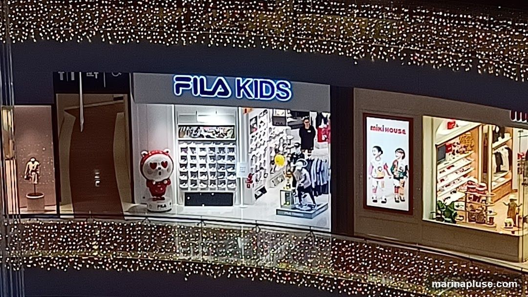 A Fila Kids store is visible with its signage above the entrance. The store displays shoes and clothing. To the left, there is a display of a mannequin wearing a hoodie. To the right, a Miki House store is visible with a poster and merchandise. The storefronts are decorated with string lights.