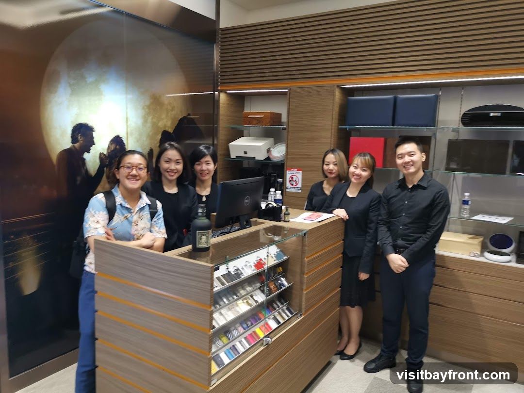A group of six people, five staff members and one customer, are posing for a photo behind a counter in a store. The staff members are all wearing black uniforms. The customer is wearing a blue and white floral shirt and glasses. The store appears to be a watch or jewelry store, with display cases filled with watches and other accessories. There is a large mural on the wall behind the counter depicting silhouetted figures against a moonlit sky.
